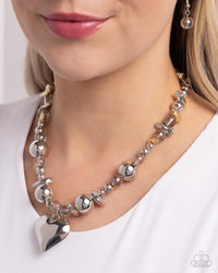 Infused along an invisible string, a collection of silver beads in varying sizes, chiseled gold pebbles, and transparent gray beads loop around the neckline for a glistening display of color. An oversized silver heart pendant swings from the center of the display for a light-hearted finish. Features an adjustable clasp closure.  Sold as one individual necklace. Includes one pair of matching earrings. 