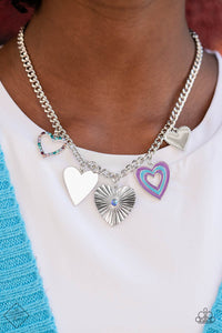 A collection of whimsical heart charms, each with its own unique features, gathers along a flat, silver curb chain. One heart is bordered in tiny silver studs, another is painted in bands of blue and purple, a third heart radiates with crimped texture and is topped with an iridescent rhinestone center, another charm radiates with its high-sheen finish, and lastly, a heart silhouette is bordered in tiny rhinestones in shades of blue and purple.