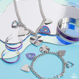 A collection of whimsical heart charms, each with its own unique features, gathers along a flat, silver curb chain. One heart is bordered in tiny silver studs, another is painted in bands of blue and purple, a third heart radiates with crimped texture and is topped with an iridescent rhinestone center, another charm radiates with its high-sheen finish, and lastly, a heart silhouette is bordered in tiny rhinestones in shades of blue and purple.